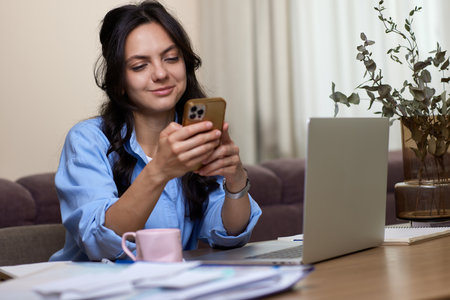 beautiful woman in blue shirt holds smart phone uses laptop in home officeの写真素材