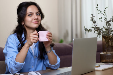 Beautiful woman in blue shirt drinking coffee working on laptop computer while sitting at home officeの写真素材