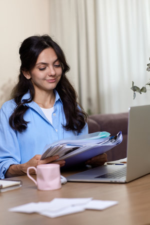 portrait of woman in blue shirt holds folders uses laptop in home officeの写真素材