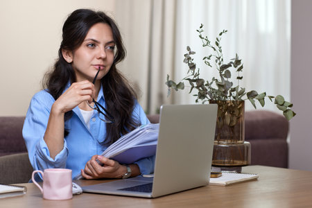 portrait of woman in blue shirt holds folders uses laptop in home officeの写真素材