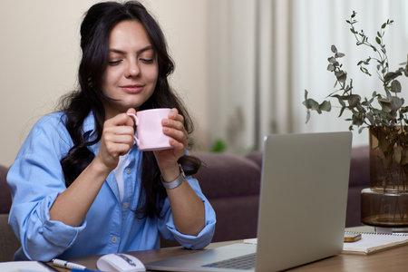 Beautiful woman in blue shirt drinking coffee working on laptop computer while sitting at home officeの写真素材