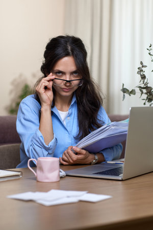 portrait of woman in blue shirt holds folders uses laptop in home officeの写真素材
