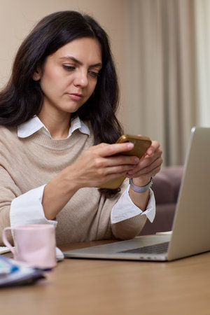 portrait of woman holds smart phone uses laptop in home officeの写真素材