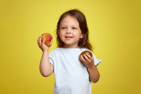 cute Caucasian little child girl holding kiwi and peach on yellow backgroundの写真素材
