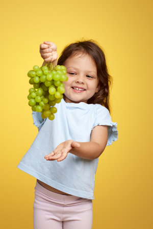 cute Caucasian little child girl holding bunch of green grapes on yellow backgroundの写真素材