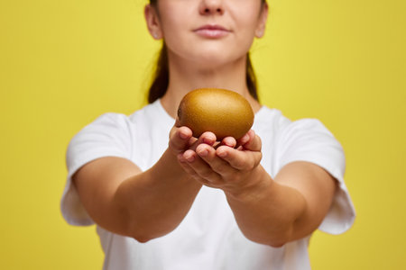 Happy woman in white t-shirt holding kiwi on studio yellow background, focus on kiwiの写真素材