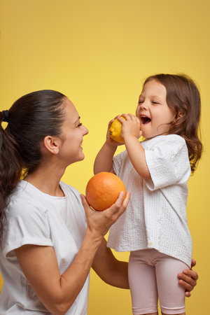 Cheerful little child girl with mother holding lemon and grapefruit on yellow background. happy family having fun togetherの写真素材