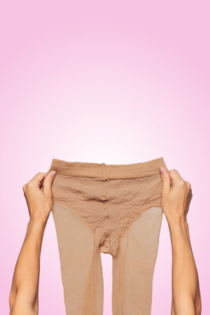 female hands holding beige nylon pantyhose on pink background.