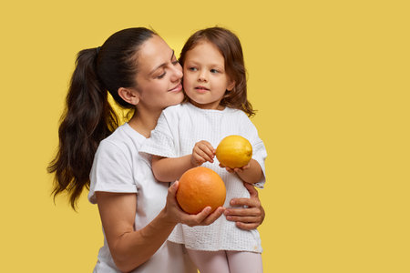 Cheerful little child girl with mother holding lemon and grapefruit on yellow background. happy family having fun togetherの写真素材
