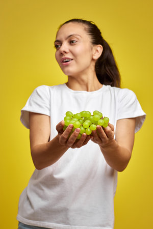 portrait of attractive Caucasian smiling woman holds grapes isolated on yellow studio background, focus on grapesの写真素材