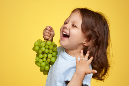 smiling Caucasian little child girl holding bunch of green grapes on yellow backgroundの写真素材
