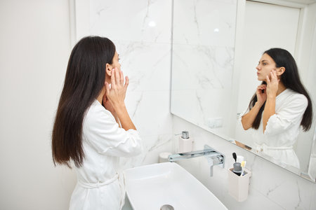Woman in a bathrobe checking her skin in the mirror as part of her morning beauty routineの写真素材