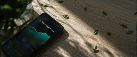Smartphone screen showing a "Screen-Free Time" graph placed on a wooden table with scattered green leaves and natural light creating soft shadows.の素材