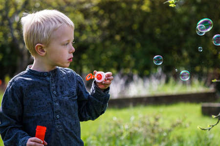 A Little boy blows soap bubbles in the garden on a summer day. He wears a blue shirt and is happy