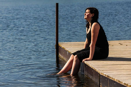 young lady sitting on the jetty and dips her feet in the cool lake water on a sunny dayの写真素材