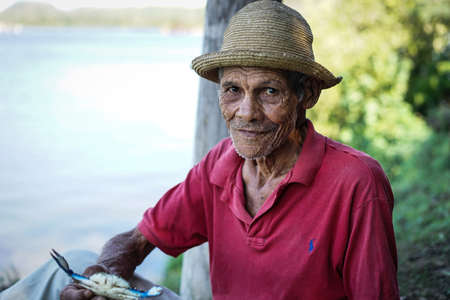 Cayo Granma, Santiago De Cuba. Cuba - January 11, 2016: old Cuban man who has caught crabs and displays the day's catch. He wears the straw hat as protection against sun rays and a red polo shirt. He is unshaven and his face is wrinkledのeditorial素材
