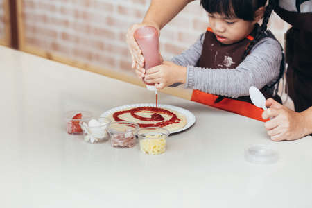 A little girl in a brown apron spread tomato sauce on the pizza doughの写真素材