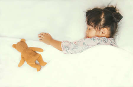 Cute Asian child sleepping with her teddy bear on white bedの写真素材