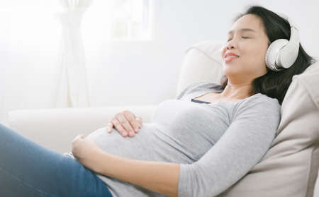 Portrait of pregnant asian woman sitting on sofa at home and listening music in headphones.の写真素材