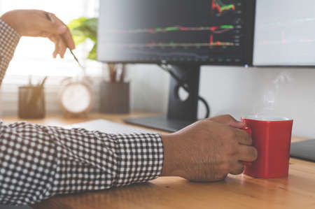 Hipster  businessman holding coffee cup and  sitting at he working place. Stock trader workplace with financial market graphs on computer monitorsの写真素材