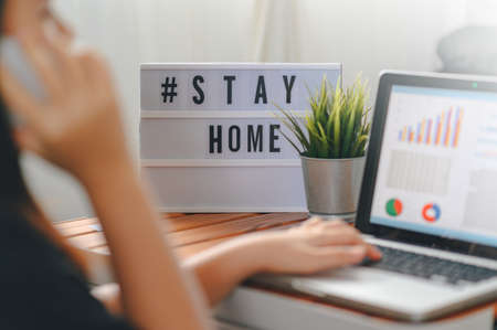 lightbox with text hashtag #STAYHOME glowing in lightand blurred woman working at home. Office worker on quarantine. Home working to avoid virus disease. Freelancer or remote worker concept.の写真素材