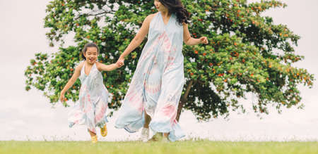 Happy family mother and daughter run on green grass at background of trees during summer sunsetの写真素材