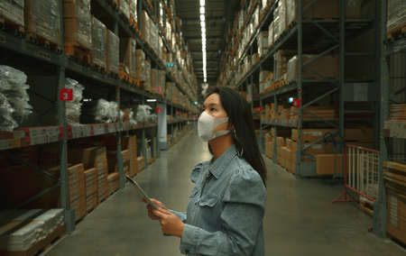 Asian business working woman wear mask using digital tablet checking stock of products in warehouseの写真素材