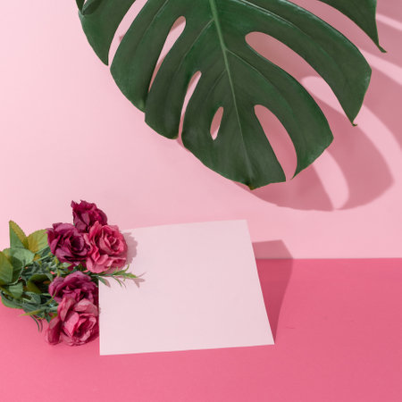Piece of blank paper next to the flower bouquet and monstera leaf on pink background. Romantic greeting card, Valentine day concept.の写真素材
