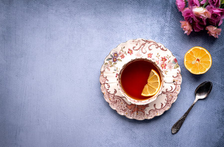 Top view of beautiful porcelaine vintage tea cup with black tea and a slice of lemon. Small rose bouquet and stylish teaspoon on the side. Gray-blue background with copy spaceの写真素材