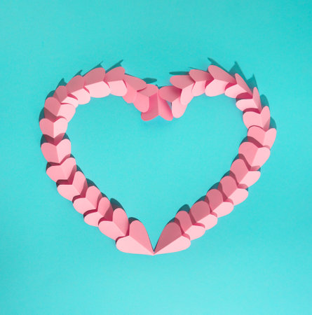 Small pink paper hearts arranged to form a big heart frame with copy space on turquoise background....