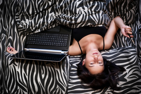 Attractive woman in bed with notebook and electronic cigarette. Top view. Cyber romance, virtual life and internet addiction conceptの写真素材