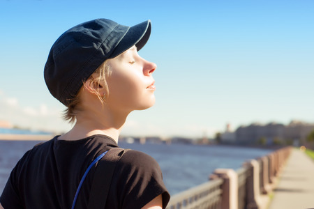 Young relaxing woman at sunny day against summer city landscape. Blue cap, eyes closed, river at backgroundの写真素材