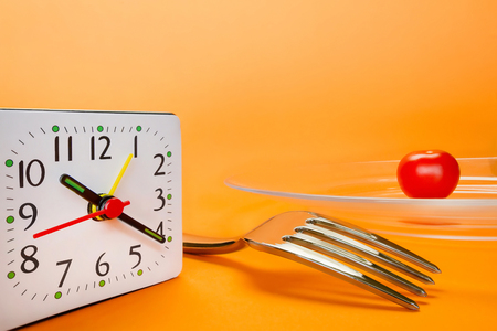 Alarm clock, fork and small cherry tomato on abstract orange background. Diet and lunch time conceptの写真素材