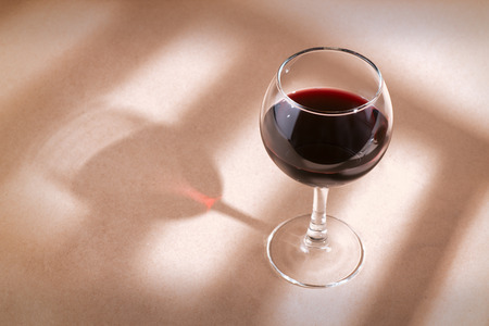Single red wine glass on the beige table with window shadow.