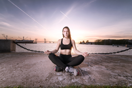 Calm athletic girl meditating on urban sea coast at sunset. Against evening skyの写真素材