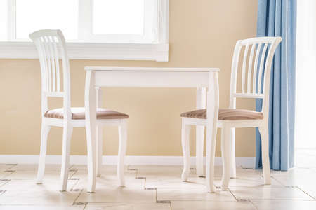 Two white chairs and white empty table by the window in a clean large room. Interior image with no...