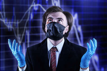 Businessman with a bruise on his face in an official suit, medical mask and gloves, raising his eyes to heaven against the blurred stock chart. Crisis, epidemic, bad luck and accident themeの写真素材