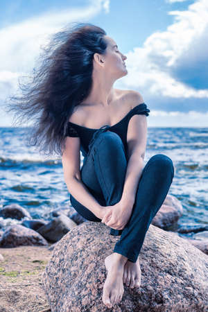 Calm brunette woman sitting on a stone by the sea with long hair flying in the wind. Blue tinted outdors portraitの写真素材