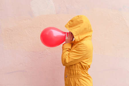 Person in yellow cloak with hood with red balloon against beige wall. Abstract conceptual portrait