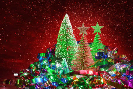 Christmas composition with tinsel, toy fir trees and sparkle defocused red background. Colorful photo, selective focus on the firの写真素材