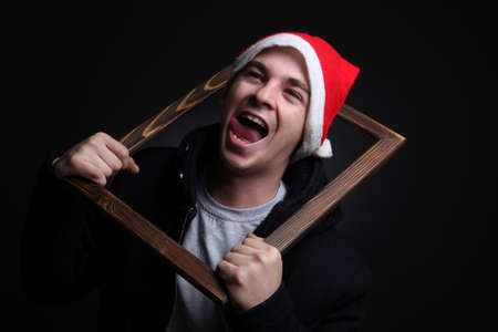 Young man in Santa hat holding wooden frame and screaming on black backgroundの写真素材