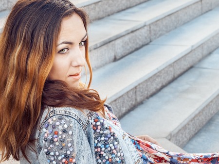 Young girl with brown hair and green eyes, dressed in long dress and jeans jacket, is sitting on the stone steps of the ladderの写真素材