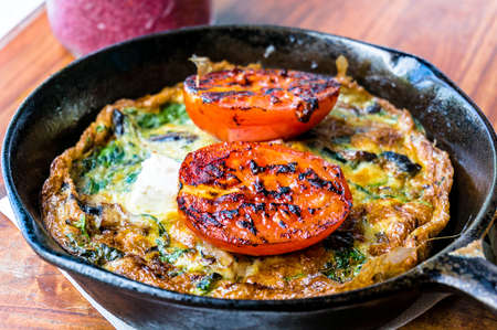 Omelette with tomato and greens on cast iron frying pan. Rustic breakfast omelette with grilled tomato and herbs. Selective focus, shallow dofの写真素材