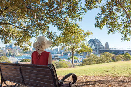 Woman sitting on a bench in a park and looking at Sydney Harbour bridgeの写真素材