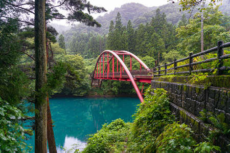 Beautiful countryside landscape with red arch bridge over blue lake surrounded by bright green...