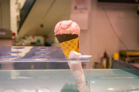 Strawberry pink ice cream in chocolate waffle cone in ice cream holder in a shop. Summer snack sale