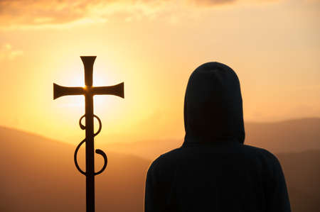 Dark silhouette of person in a hood and Christian cross against sun on the background. Abstract religious backgroundの写真素材