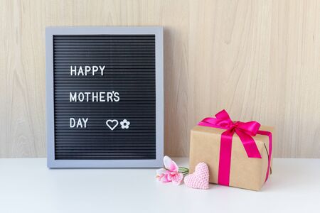 Decorative sign with the message: Happy Mother's Day; Beside have a flower, a crochet heart and a beautiful gift wrapped in a pink ribbon. Wooden background and white base.の写真素材