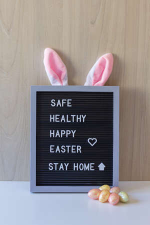 Decorative sign with rabbit ears and colored eggs in front with the easter message: Safe, healthy, happy easter, stay home.の写真素材