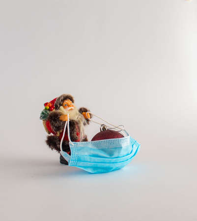 Virus during the holidays. Santa pulls a decorative ball in a face protective mask on white...